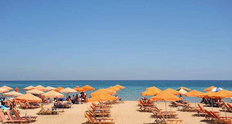Reihen von orangefarbenen Sonnenliegen und Sonnenschirmen mit Blick auf ein ruhiges blaues Meer an einem Sandstrand.