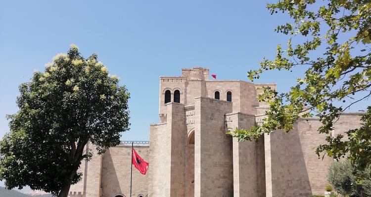 Los muros de piedra clara del Castillo de Krujë y la bandera albanesa enmarcados por un árbol frondoso contra un cielo despejado.