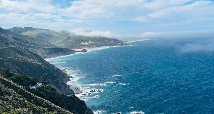 Sweeping view along the rugged Big Sur coastline where green hills drop to the crashing blue Pacific beneath patches of coastal fog.