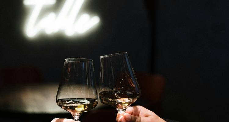 Dos manos chocando copas de vino en un bar con poca luz y un fondo con un letrero de neón de estilo.