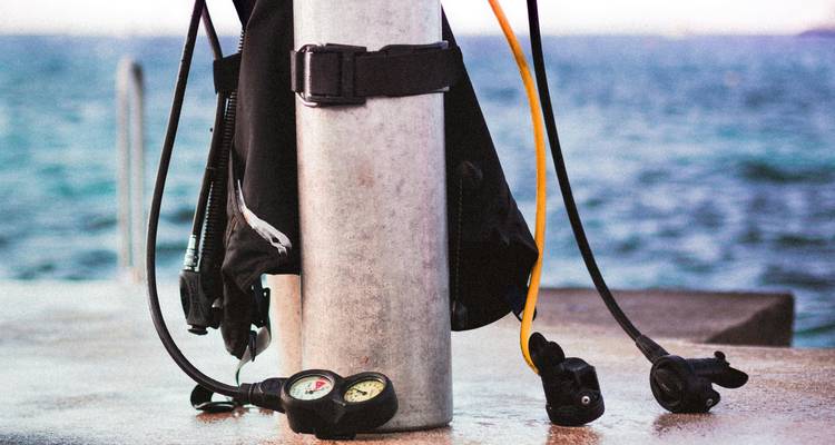 Un tanque de buceo con reguladores y medidores adjuntos está de pie en un muelle con el mar azul de fondo.