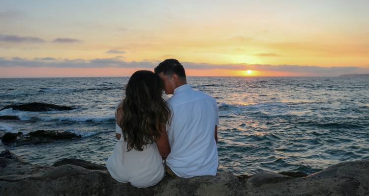 Una pareja está sentada en las rocas mirando cómo el sol se pone sobre un mar tranquilo y ondulante.