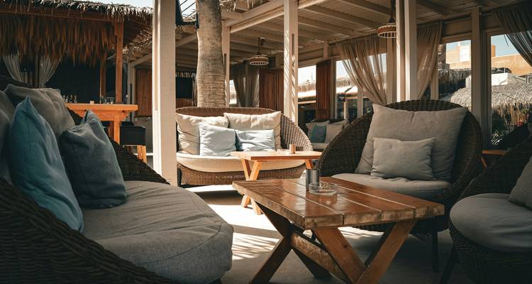 Área de descanso al aire libre con estilo, con sofás de mimbre, cojines y una mesita de café de madera bajo la luz del sol.