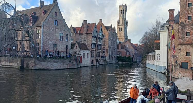 Canal panorama with boats of tourists and Bruges skyline under mixed clouds.
