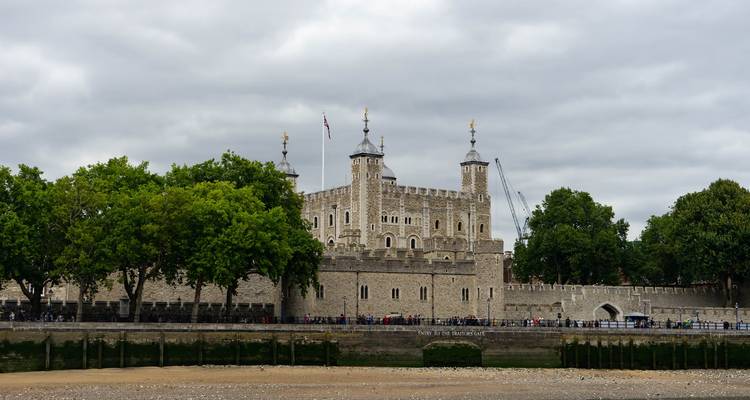 Tower of London viewed across the Thames with tree-lined embankment under a gray sky