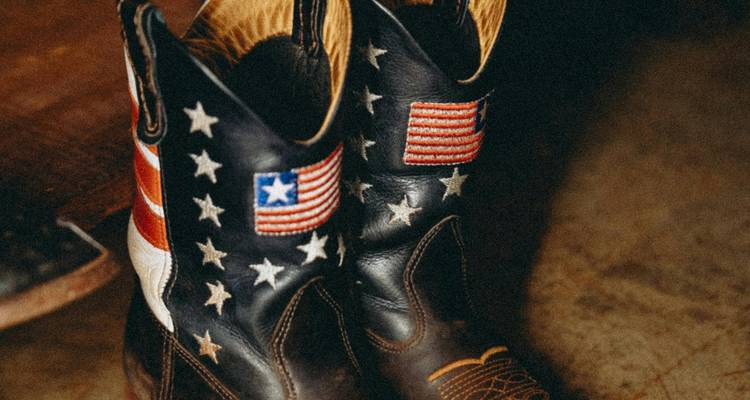 Paire de bottes de cowboy en cuir usé décorées de patchs du drapeau américain et d'étoiles à l'intérieur sur un sol rustique