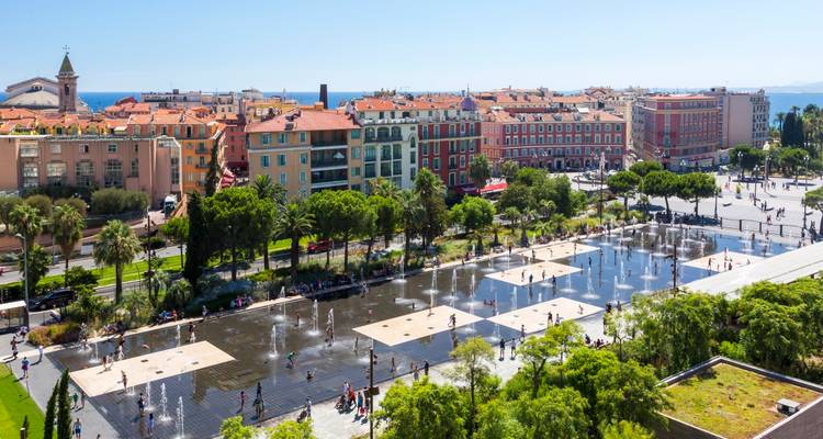 Grand parc urbain avec des jets d'eau ludiques et des personnes marchant sur des fontaines en damier, des bâtiments historiques en arrière-plan.