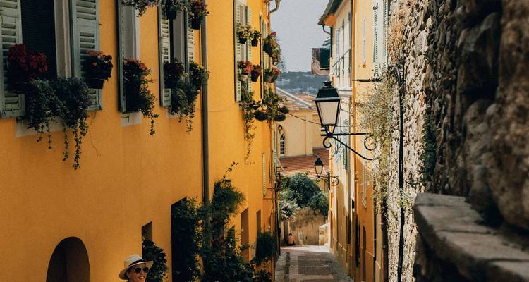 Lebendige gelbe Gasse gefüllt mit Blumenkästen; Reisender in Sonnenhut darunter gehend.