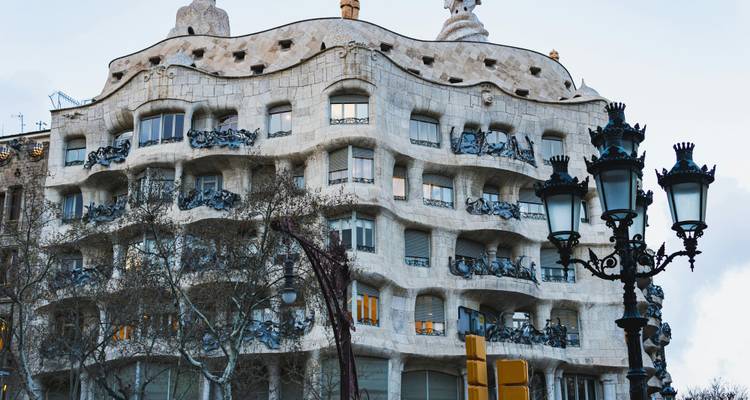 Wellenförmige Steinfassade von Gaudís Casa Milà mit Winterbäumen und ornamentalen Straßenlaternen im Vordergrund