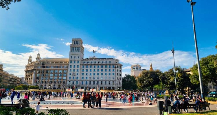 Lebhafter Plaça de Catalunya mit Brunnen, Tauben, Menschenmengen und historischen Gebäuden unter einem leuchtend blauen Himmel