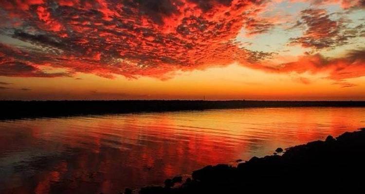 Vivid red and orange sunset clouds reflected on calm water with dark shoreline silhouette.