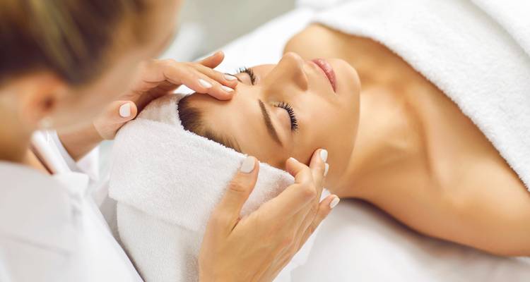Spa therapist gently massages a woman's forehead as she relaxes under a white towel.