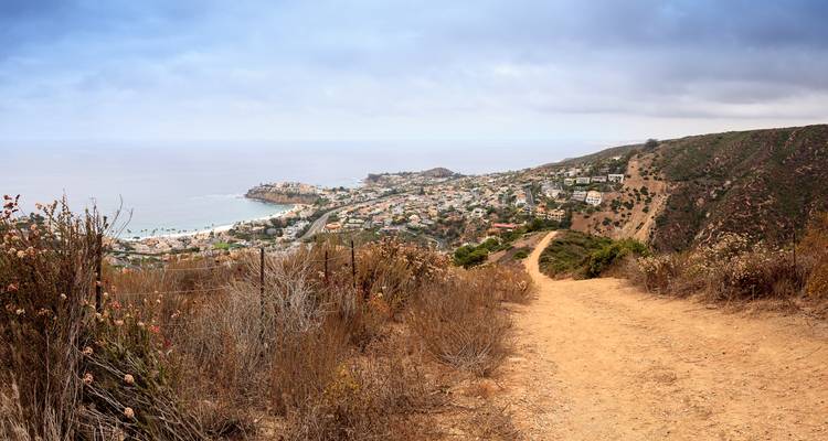 Dusty hiking trail on a dry hillside overlooking a coastal town and expansive ocean under partly cloudy sky.