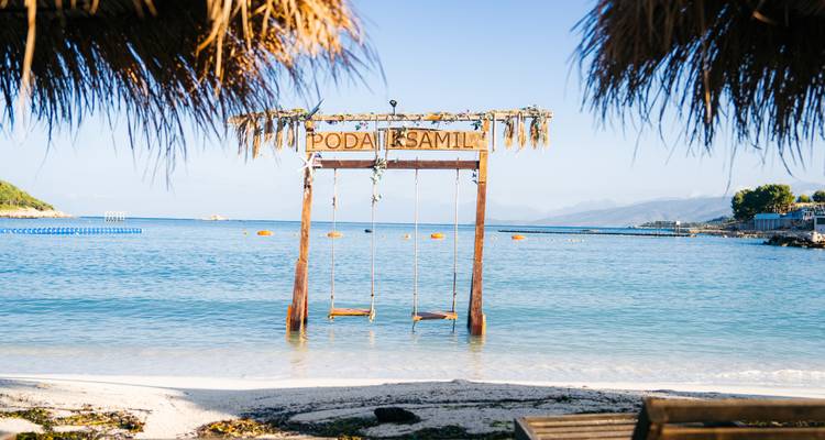 Dekorativer Holzschaukelstand in seichtem türkisfarbenem Wasser, umrahmt von palmgedeckten Sonnenschirmen, Schild mit Aufschrift "Poda Ksamil"
