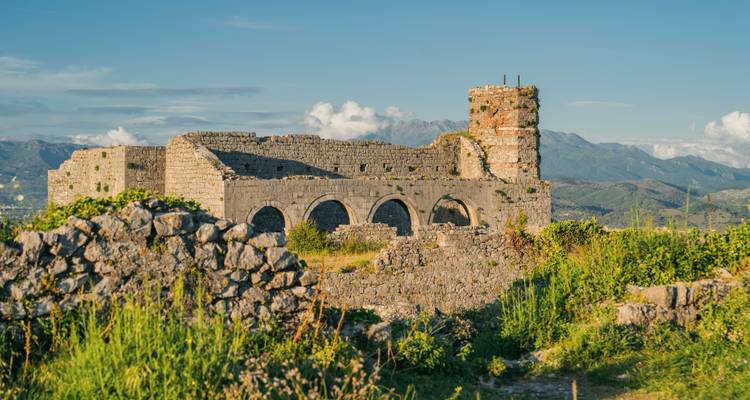 Sonnendurchflutete Ruinen eines Steinschlossturms und Bögen, umgeben von grünem Gras mit Bergen im Hintergrund