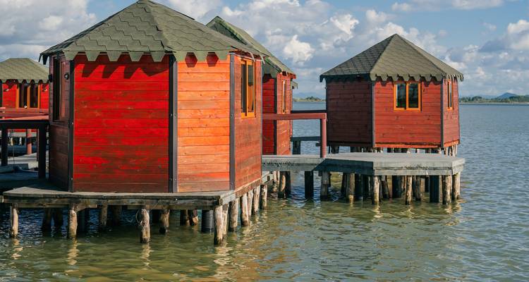 Gruppe roter Holzbungalows auf Stelzen über ruhigem Lagunenwasser unter teilweise bewölktem Himmel