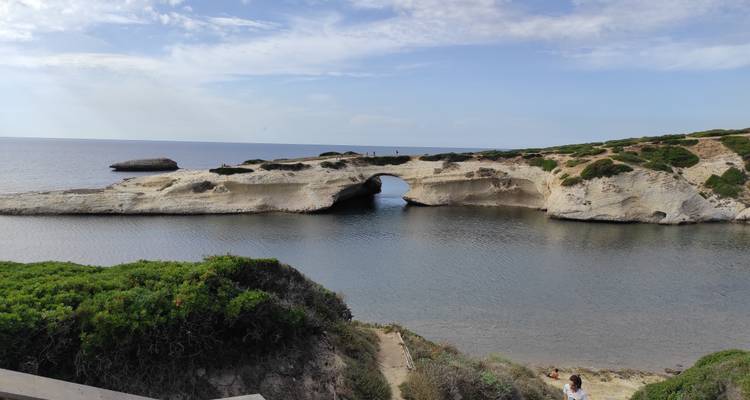 Arche en pierre blanche naturelle s'étendant dans une mer calme à côté de falaises basses couvertes de buissons et d'un horizon bleu.