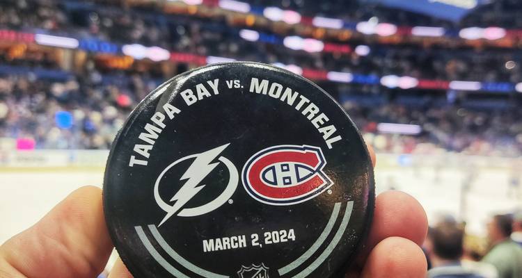 Gros plan d'un disque de hockey lisant « Tampa Bay vs. Montréal » tenu à l'intérieur d'une arena de hockey sur glace avec foule et patinoire floues en arrière-plan.