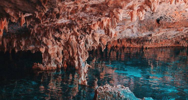 Piscine souterraine colorée avec des stalactites orange dentelées se reflétant dans l'eau bleue.