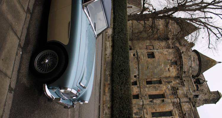 Voiture de sport bleue vintage garée devant un ancien château écossais par une journée nuageuse.