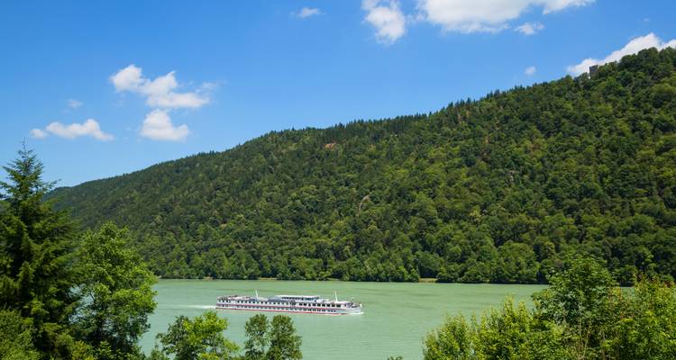 Un barco blanco de crucero fluvial viaja por un amplio río verde bordeado por exuberantes colinas boscosas bajo un cielo azul brillante con nubes dispersas.