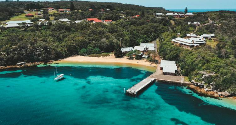 Luchtfoto van turquoise baai met steiger, zeilboot en bebost nederzetting op Maria Island.