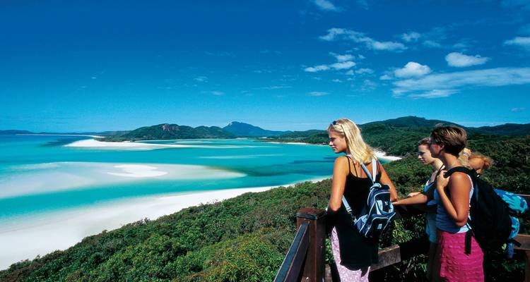 Group of backpackers at a wooden lookout admire the turquoise swirling sands and islands of Hill Inlet in the Whitsunday Islands under clear skies.