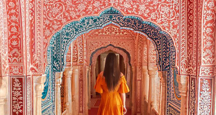A woman in a flowing orange dress walks through the ornate pink and blue arches of Jaipur’s Patrika Gate.
