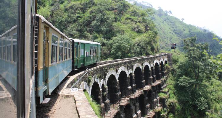 A vintage narrow-gauge train crosses a high stone viaduct through lush green Himalayan foothills.