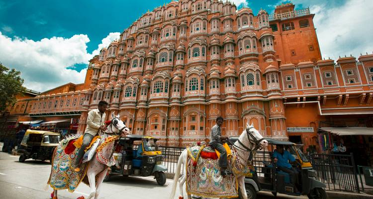 Colourfully dressed riders on decorated horses pass the elaborate pink façade of Jaipur’s Hawa Mahal under a bright sky.