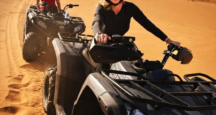 Twee rijders rijden op quad bikes over goudkleurig woestijnzand en laten sporen achter zich.