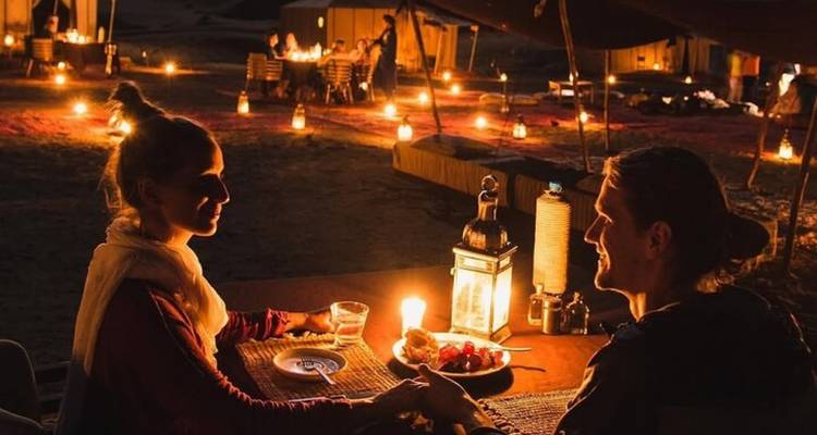 Een stel geniet van een kaarsenlicht diner in een woestijnkamp omgeven door gloeiende kaarsen.