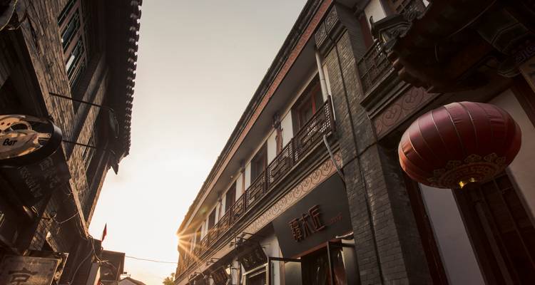 Rue étroite bordée de bâtiments traditionnels et d'une lanterne rouge, avec un reflet de soleil apparaissant au crépuscule.