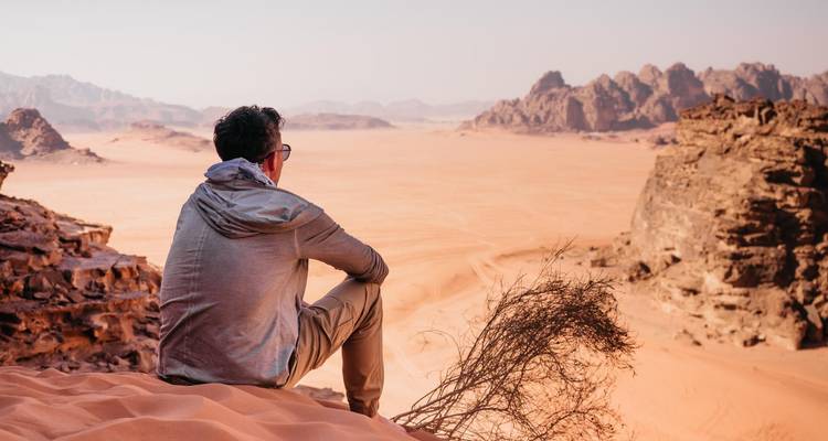 Man sitting on a rocky ledge overlooking an expansive desert of golden sand and rugged mountains