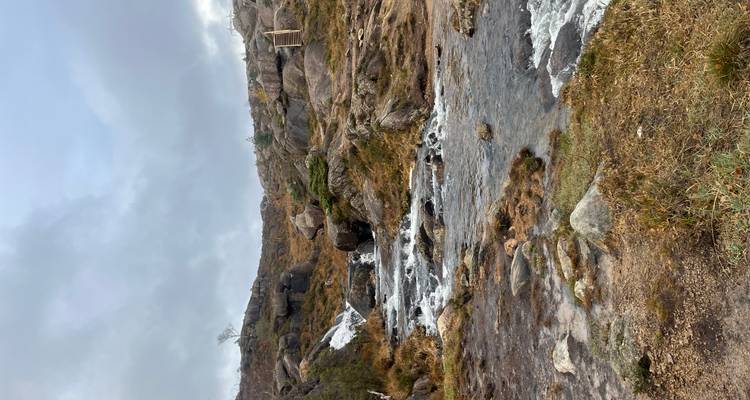 Ruisseau rocheux peu profond avec de petites cascades s'écoulant à travers un terrain aride semblable à la toundra sous un ciel gris.