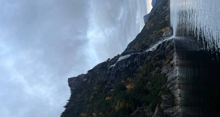 Une haute cascade se déversant le long d'une falaise boisée dans un fjord au crépuscule sous des nuages chargés.