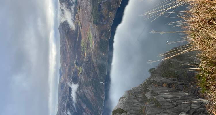 Vue panoramique élevée surplombant une paroi rocheuse abrupte donnant sur un fjord brumeux et des champs de plateau vallonnés.