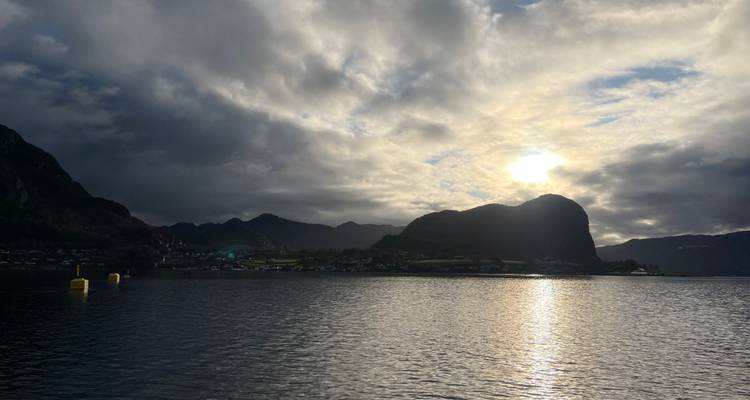 Le soleil brillant à travers des nuages dramatiques au-dessus de montagnes sombres en silhouette et d'un fjord aux eaux réfléchissantes.