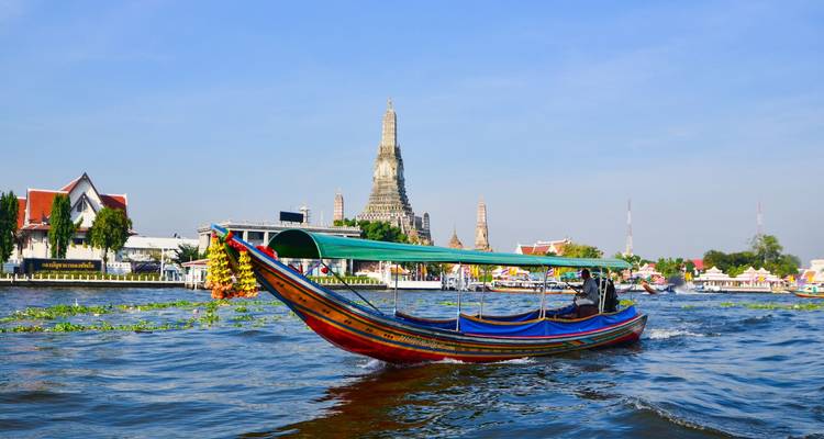 Un bateau à queue longue navigue rapidement le long de la rivière Chao Phraya avec le Wat Arun se dressant en arrière-plan sous un ciel bleu.