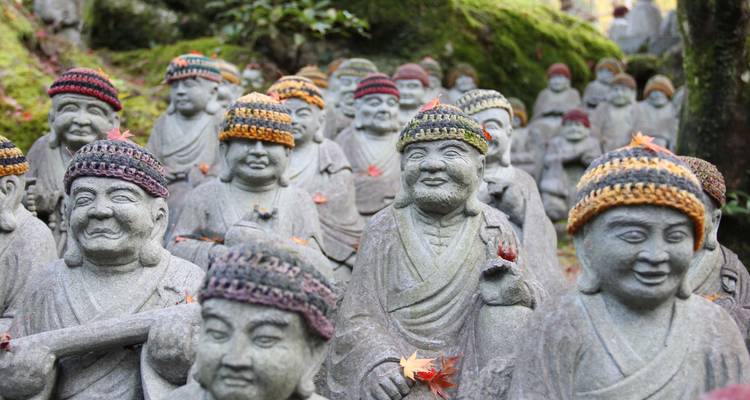 Grupo de estatuas budistas de piedra con gorros de lana tejida de colores en un bosque cubierto de musgo.