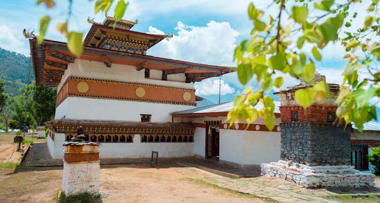 Kleiner weiß getünchter bhutanischer Tempel mit farbigem Dach und Gebetsmühlen, umrahmt von grünen Blättern