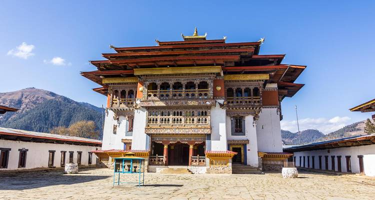 Prachtvolles bhutanisches Dzong mit weiß getünchten Mauern und reich bemalten Holzfenstern vor einem klaren blauen Alpenhimmel.