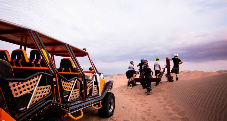 Adventure group with boards prepares to sandboard beside an orange dune buggy on vast desert dunes.