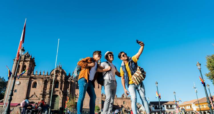 Three young travellers take a selfie against the blue sky in Cusco’s main square with colonial buildings behind.