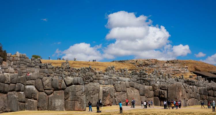 Massive stone walls of Sacsayhuaman ruins with tourists exploring the grassy esplanade under a bright blue sky.