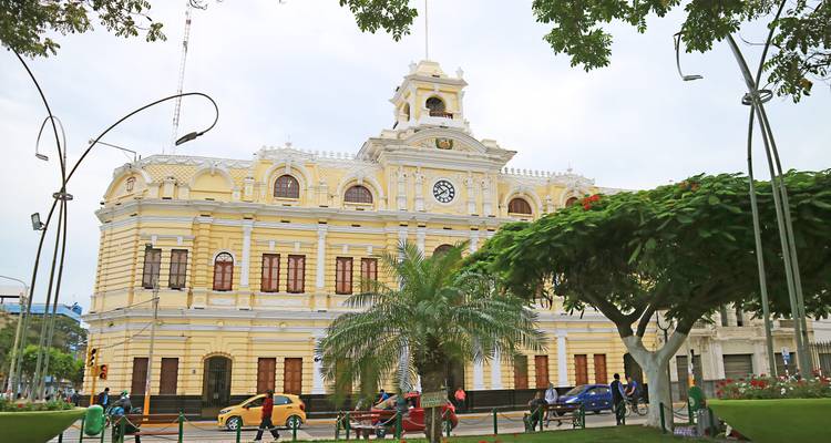 Yellow colonial civic building with a central clock tower bordered by palm trees and locals relaxing on benches.