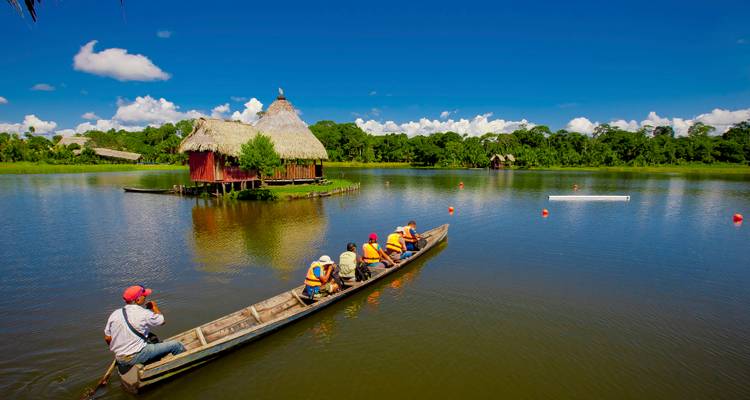 Canoe carrying life-jacketed visitors on a calm jungle lagoon with thatched huts and lush rainforest onshore.
