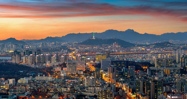 Expansive modern city skyline glowing at dusk beneath a colorful sky and distant hills.