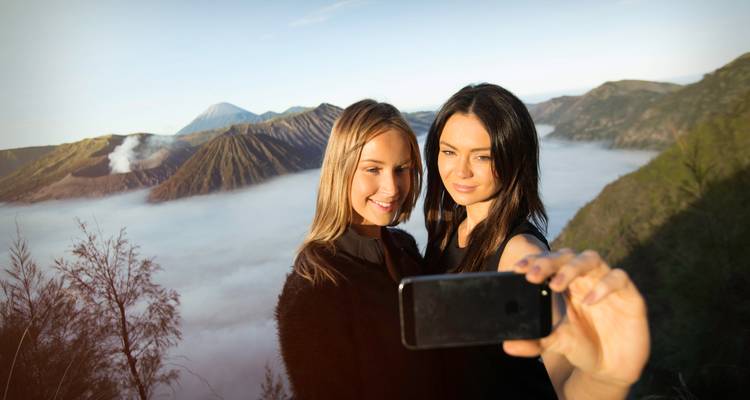Deux femmes prennent un selfie sur une crête au-dessus de nuages bas avec le cratère du Mont Bromo derrière elles.
