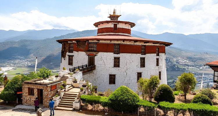 Round, white-stone Ta Dzong museum overlooking a lush valley under bright skies.